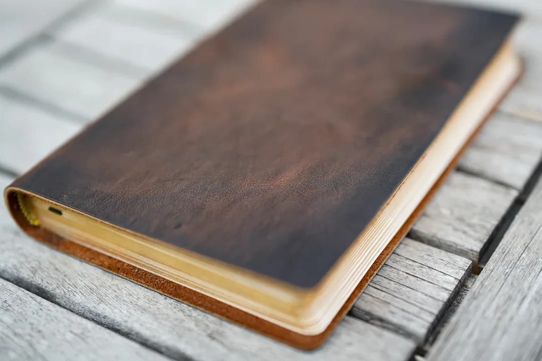 A full-grain brown leather Bible with gilded page edges, resting on weathered gray wood.